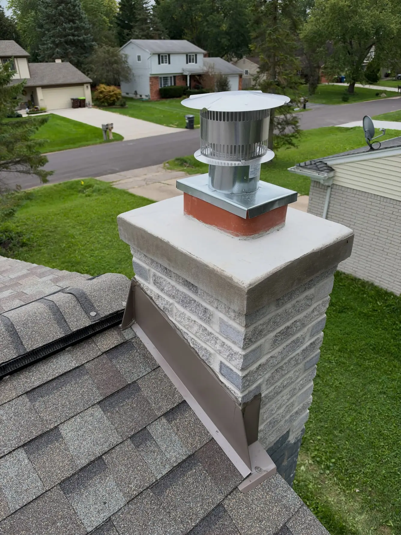 Light gray brick chimney with a new concrete crown, a stainless steel cap on the flue and metal flashing where it meets the shingle roof