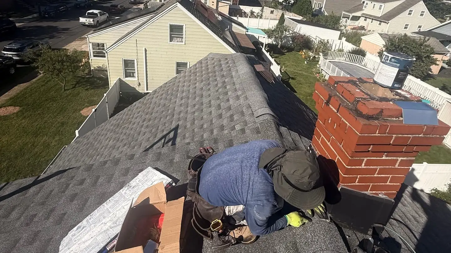 Roofer working at the base of a brick chimney on a shingle roof, with neighboring houses and yards below