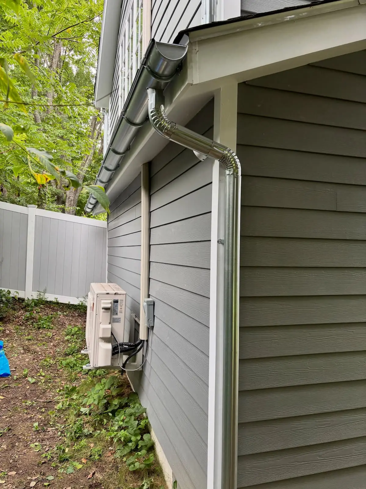 White gutter and downspout running down the corner of a house with gray lap siding, beside a wall mounted heat pump unit