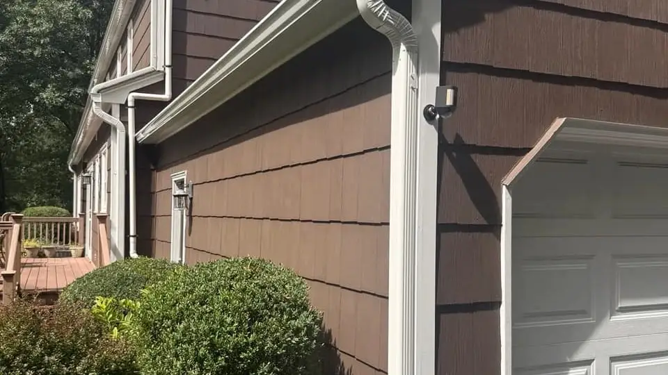 White gutter along the eave of a house with brown shake siding, and a downspout running down beside the garage door
