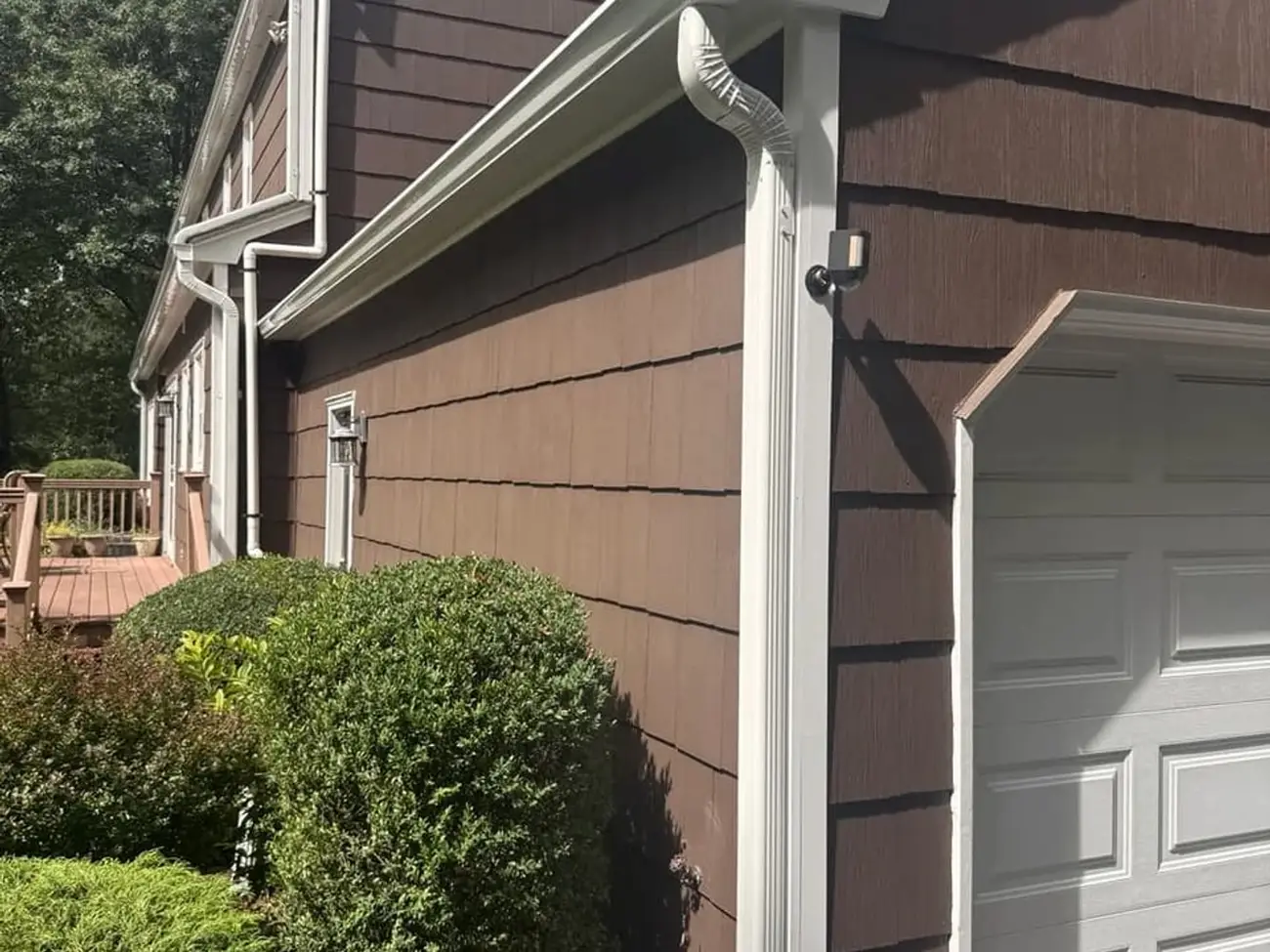 White seamless gutter running along an eave into an elbow and downspout, fixed against shake siding beside a garage