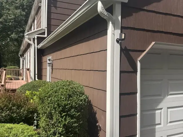 White seamless gutter running along an eave into an elbow and downspout, fixed against shake siding beside a garage