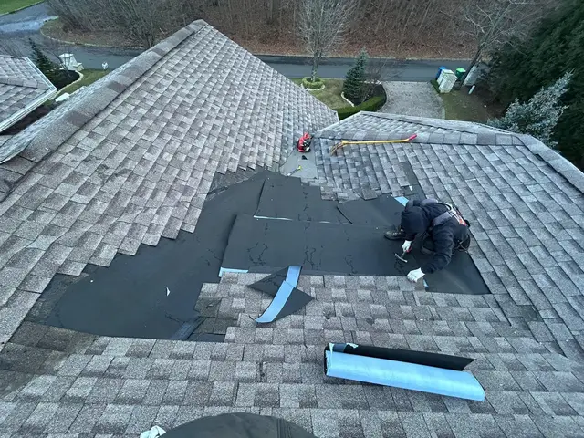 Roofer nailing down a patched section of a shingle roof, with the old shingles cut back around the repair and a roll of underlayment beside it