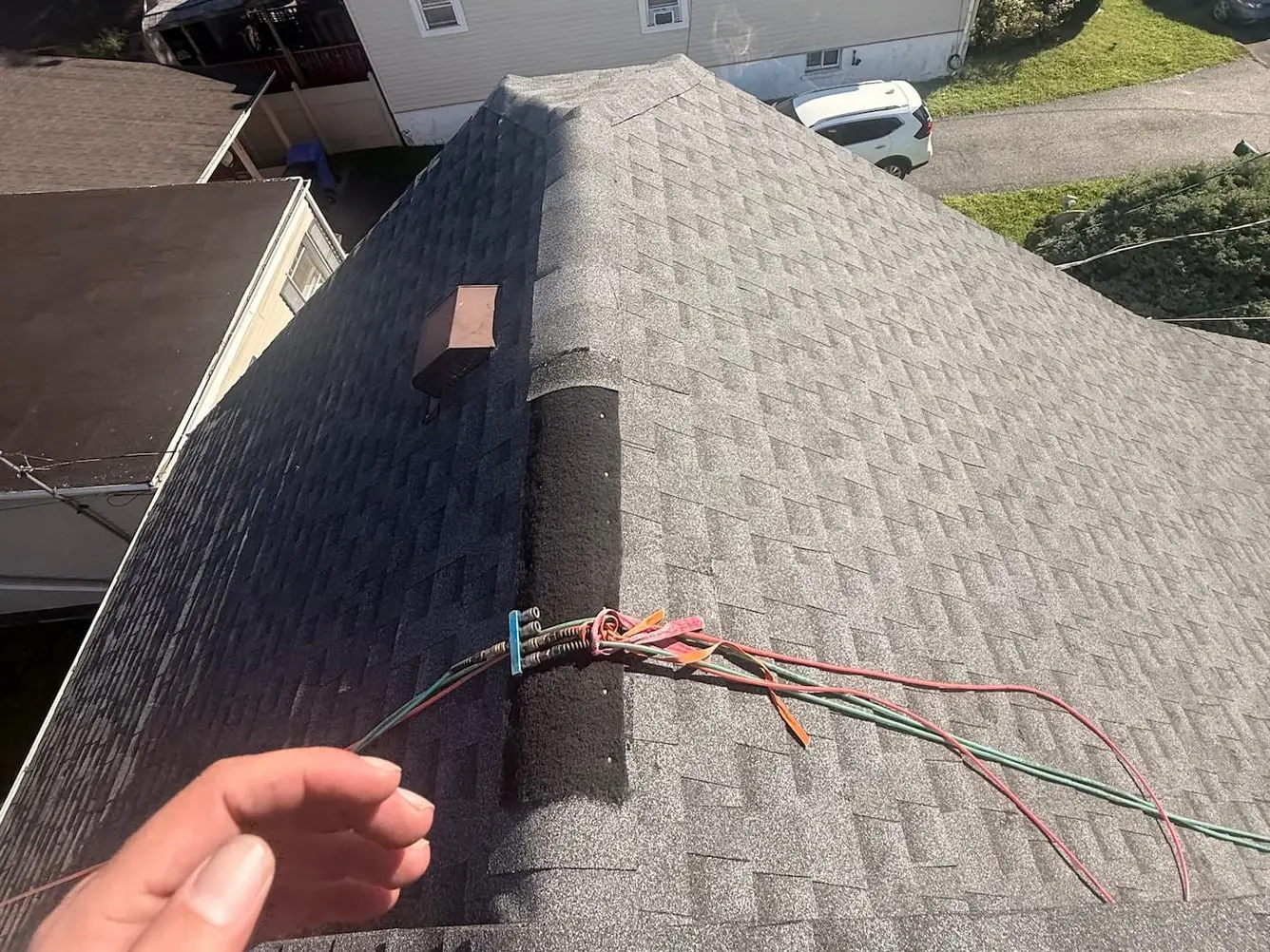 Hand at the ridge of a shingle roof lifting a bundle of loose low voltage wires that run across the shingles beside a vent