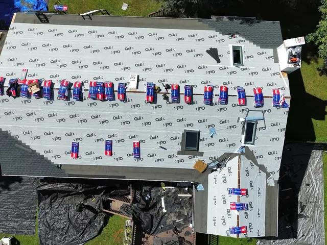 Overhead view of a fully stripped roof covered in synthetic underlayment, with around twenty shingle bundles staged in a row along the ridge and tarps below catching the torn off shingles