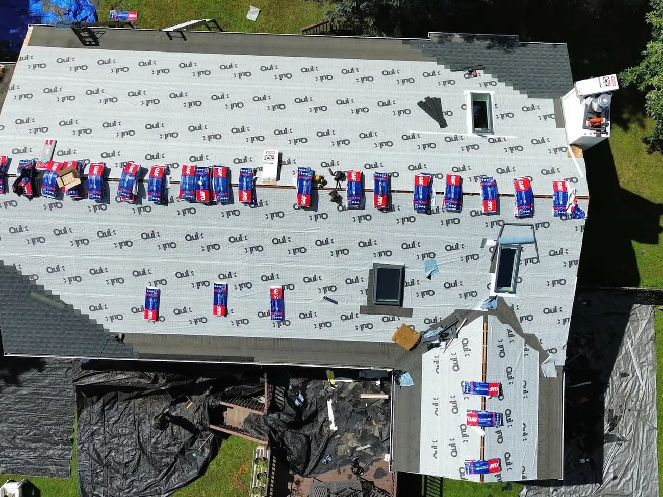 Overhead view of a fully stripped roof covered in synthetic underlayment, with around twenty shingle bundles staged in a row along the ridge and tarps below catching the torn off shingles