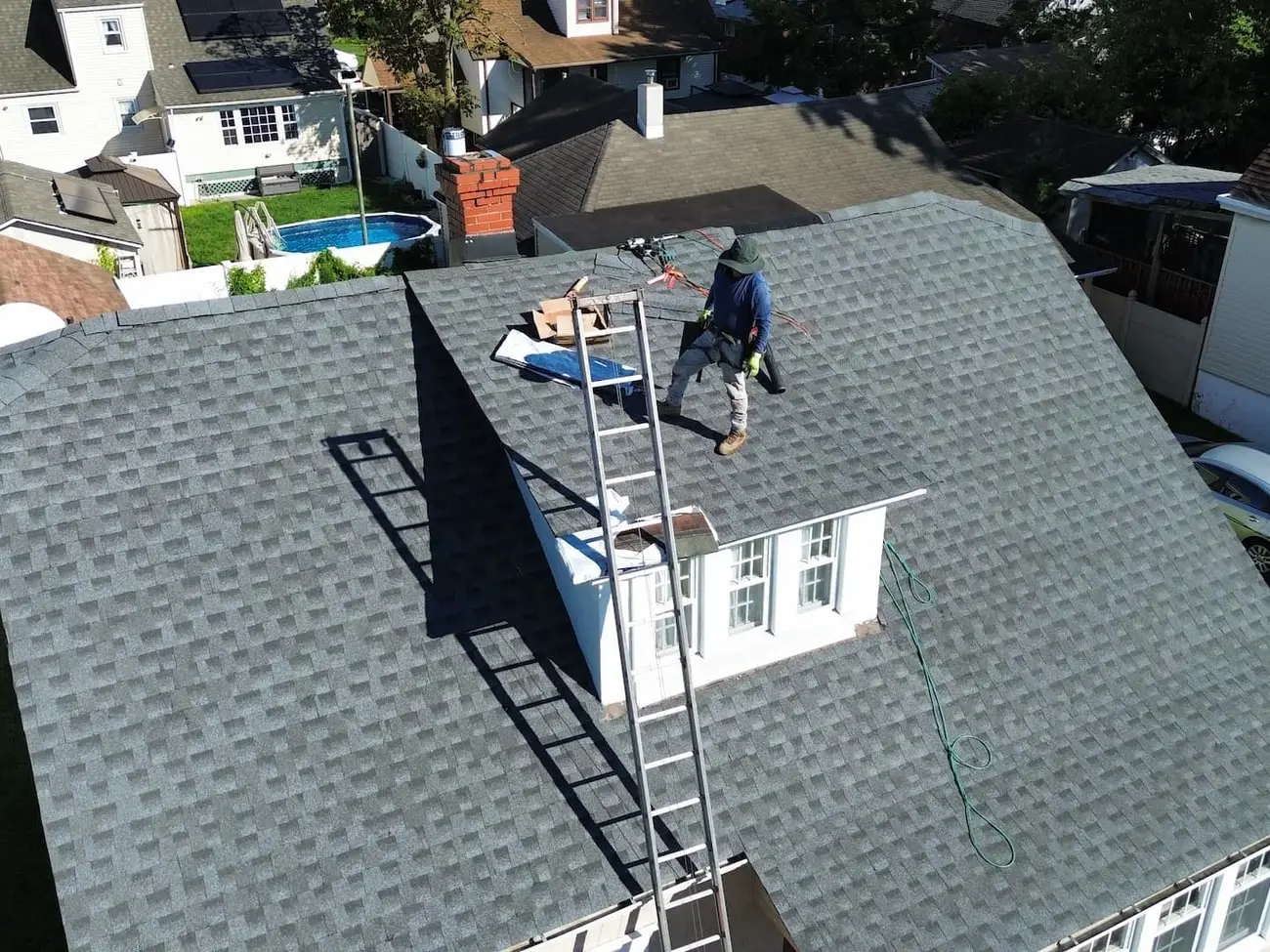Crew member standing on a finished shingle roof with a ladder set to the ridge, surrounded by neighbouring houses and driveways