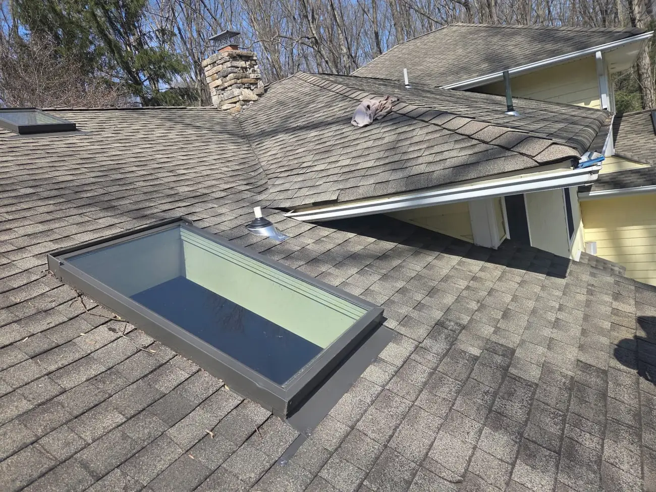 Skylight set into an asphalt shingle roof with metal flashing around the frame and a stone chimney behind it