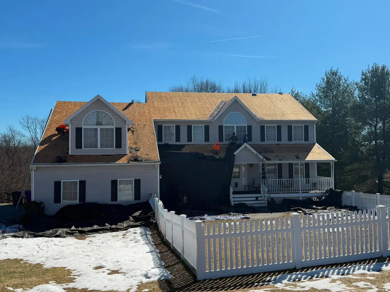 Roofing crew stripping old shingles down to the wood deck on a two-story house, with snow on the lawn