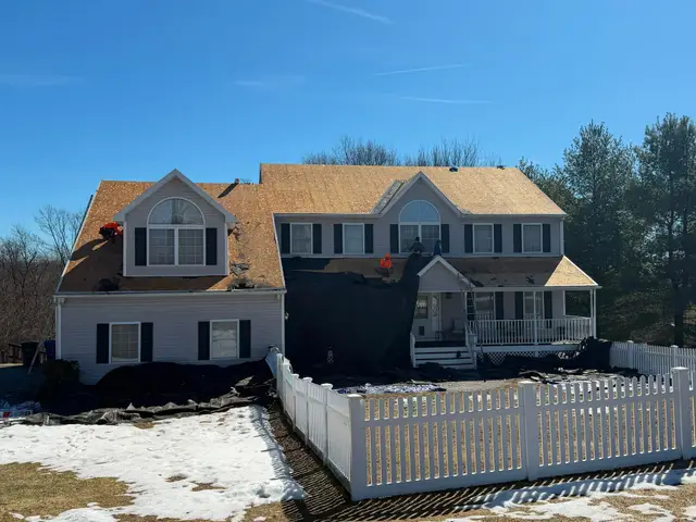 Roofing crew stripping old shingles down to the wood deck on a two-story house, with snow on the lawn